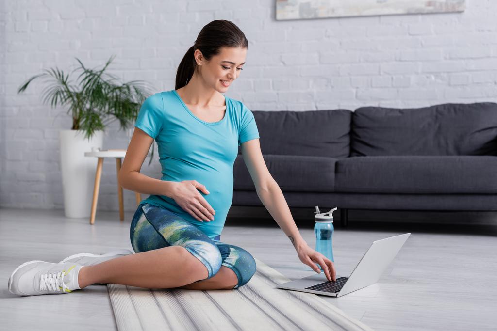 cheerful pregnant woman looking at laptop and sitting on fitness mat  - Photo, Image