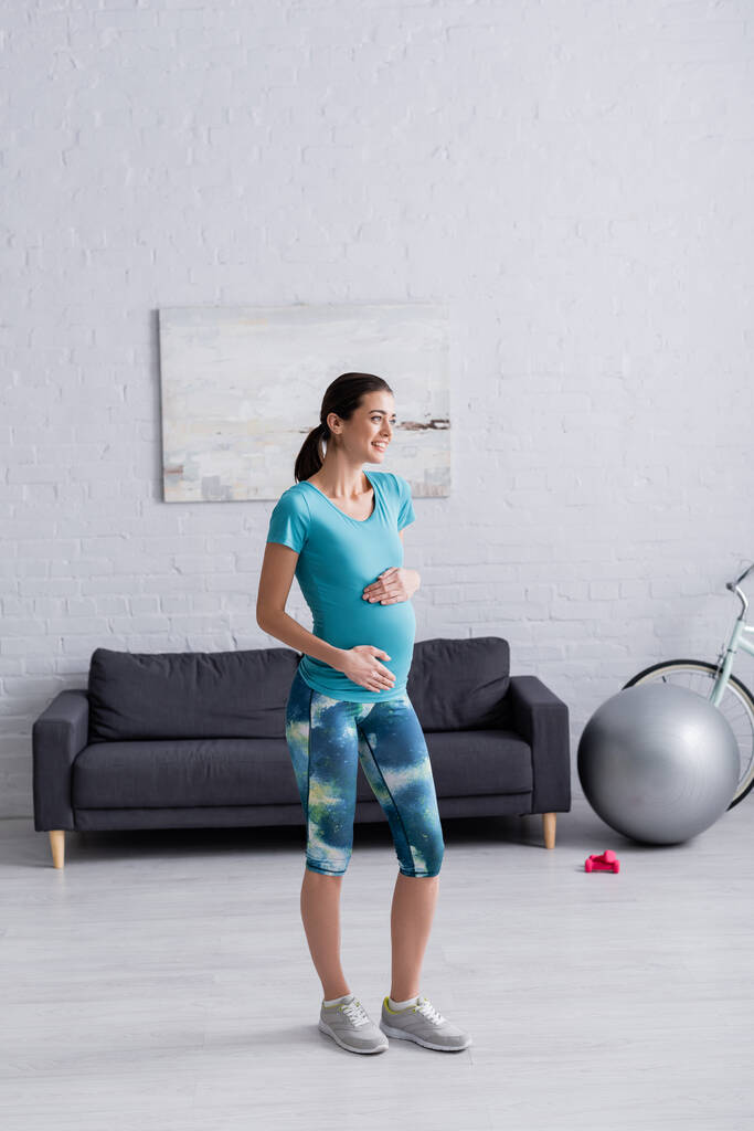 full length of cheerful pregnant woman touching belly in living room  - Photo, Image