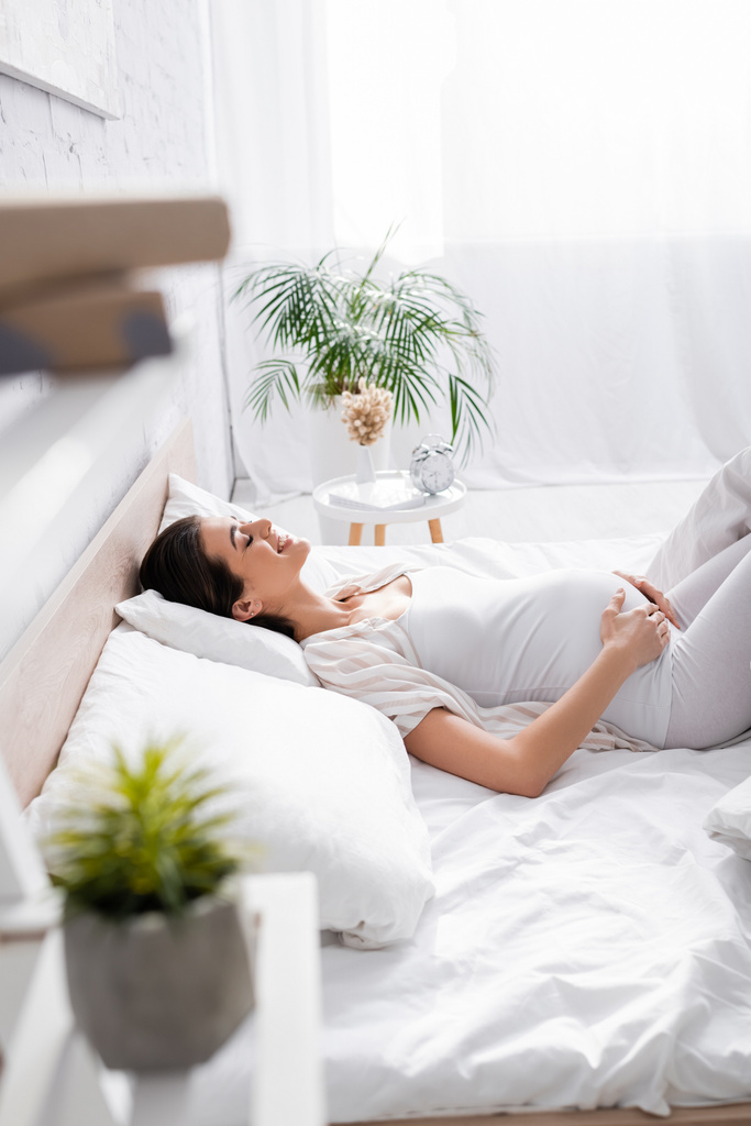 вид сбоку счастливой молодой беременной женщины, трогающей животик, лежа в спальне - Фото, изображение