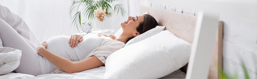 вид сбоку счастливой молодой беременной женщины, трогающей животик, лежа в спальне, баннер  - Фото, изображение