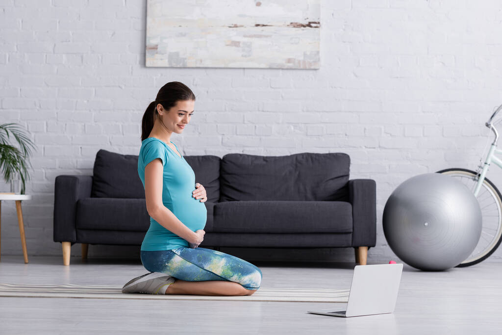 mulher grávida feliz em sportswear trabalhando perto de laptop no tapete de fitness - Foto, Imagem