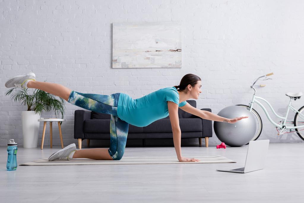 vue latérale de gaie femme enceinte en vêtements de sport travaillant près d'un ordinateur portable sur tapis de fitness - Photo, image