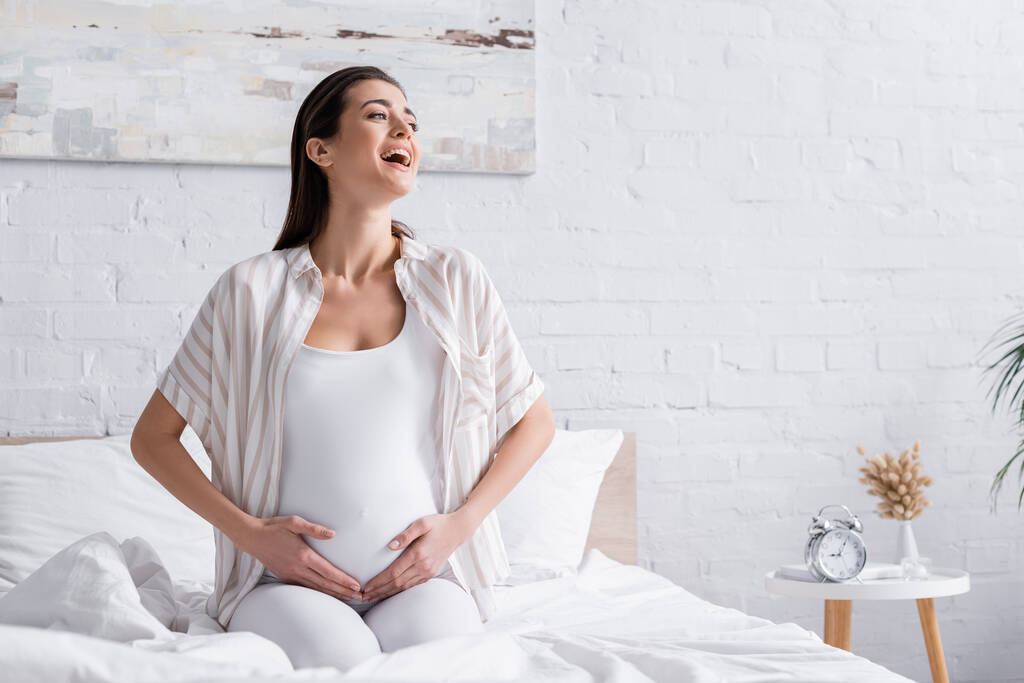 glücklich schwangere Frau lacht und berührt Bauch im Schlafzimmer - Foto, Bild