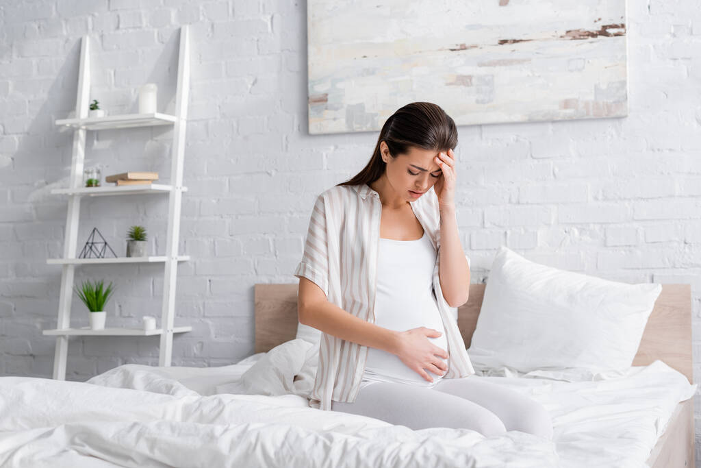 vyčerpaná těhotná žena dívá na břicho v ložnici - Fotografie, Obrázek