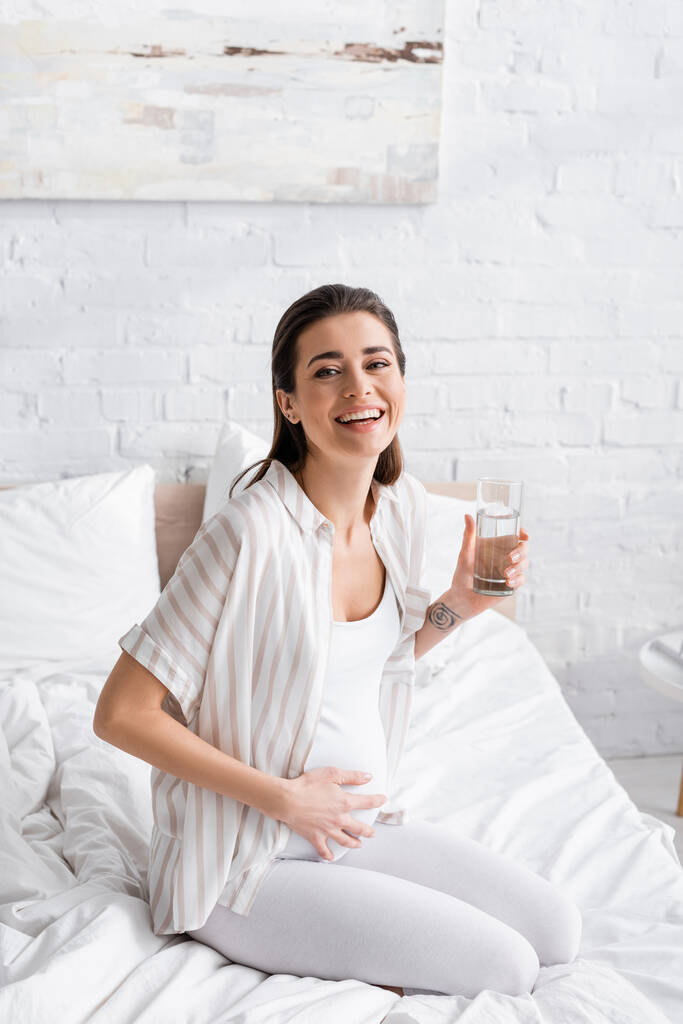 šťastná těhotná žena drží sklenici vody v ložnici - Fotografie, Obrázek