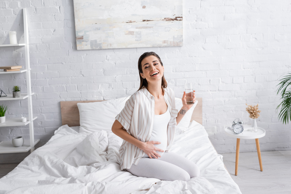 alegre embarazada sosteniendo vaso de agua en el dormitorio - Foto, imagen