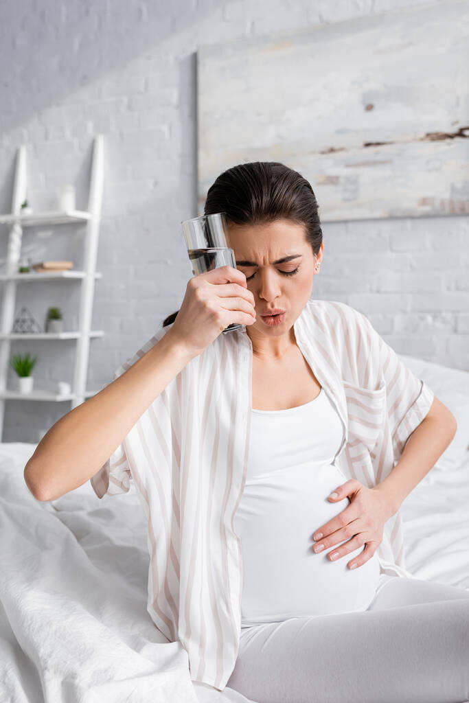 νεαρή έγκυος γυναίκα αισθάνεται κράμπα και κρατώντας ένα ποτήρι νερό - Φωτογραφία, εικόνα