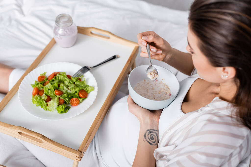 vista de ángulo alto de la mujer embarazada sosteniendo tazón con harina de avena cerca de bandeja con ensalada - Foto, Imagen