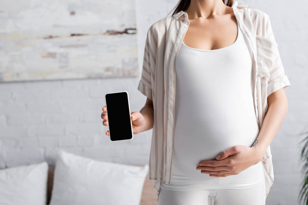 vista parcial de una joven embarazada sosteniendo un smartphone con pantalla en blanco - Foto, imagen
