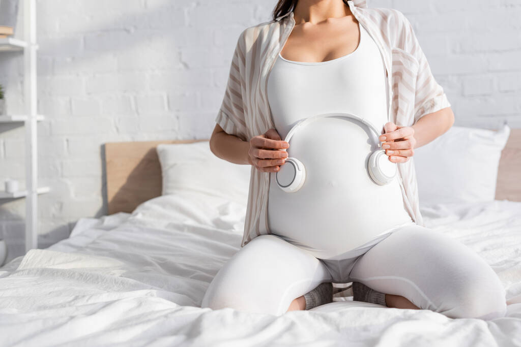 przycięty widok młodej kobiety w ciąży trzyma słuchawki w pobliżu brzucha - Zdjęcie, obraz