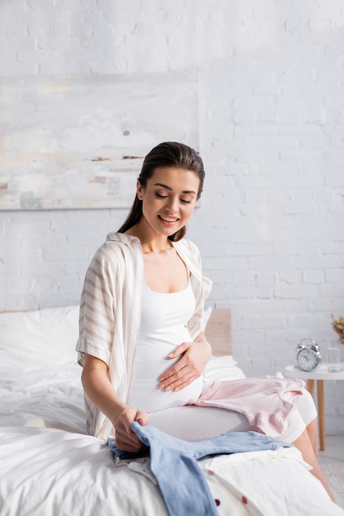 happy pregnant woman looking at baby clothes on bed - Photo, Image