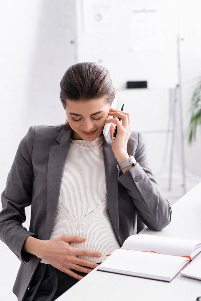 счастливая беременная деловая женщина разговаривает на смартфоне и трогает живот - Фото, изображение