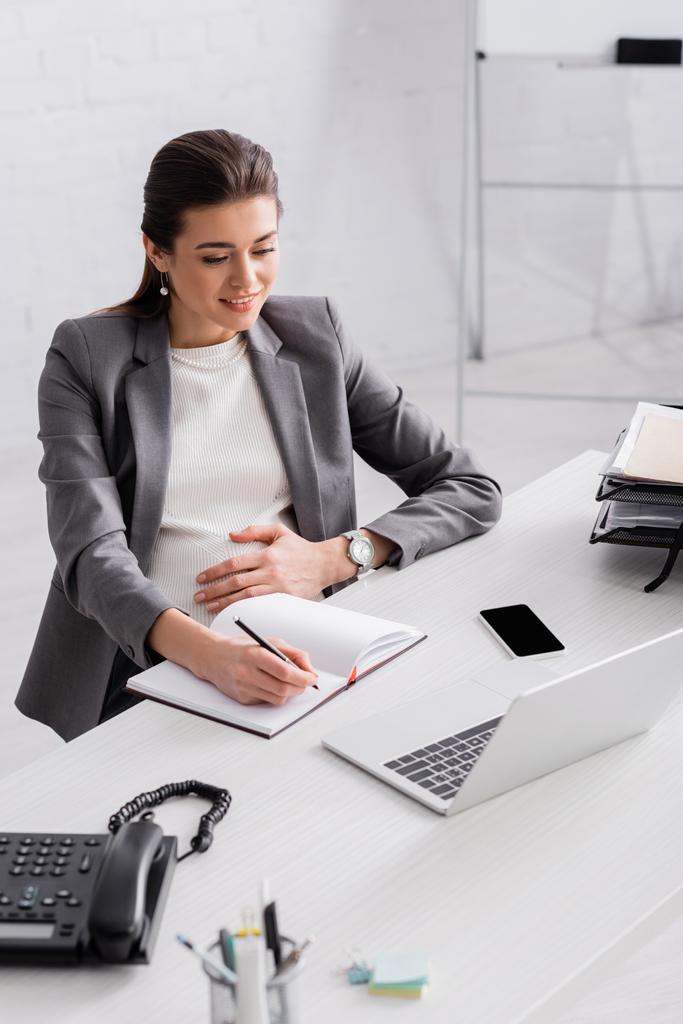χαμογελώντας έγκυος επιχειρηματίας που γράφει στο σημειωματάριο κοντά gadgets στο γραφείο - Φωτογραφία, εικόνα