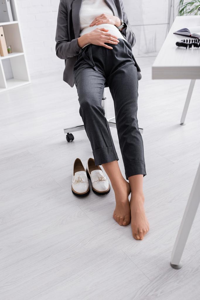 částečný pohled na těhotnou ženu bez bot sedící na židli v kanceláři - Fotografie, Obrázek