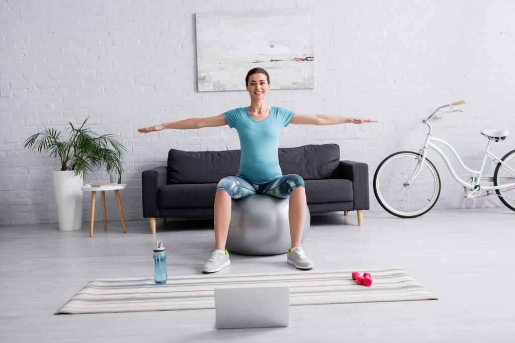 fröhliche Schwangere in Sportbekleidung beim Turnen auf Fitnessball neben Laptop im Wohnzimmer  - Foto, Bild
