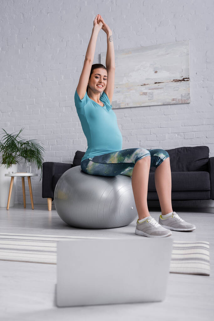 mulher grávida positiva com as mãos levantadas exercitando na bola de fitness e olhando para laptop na sala de estar  - Foto, Imagem
