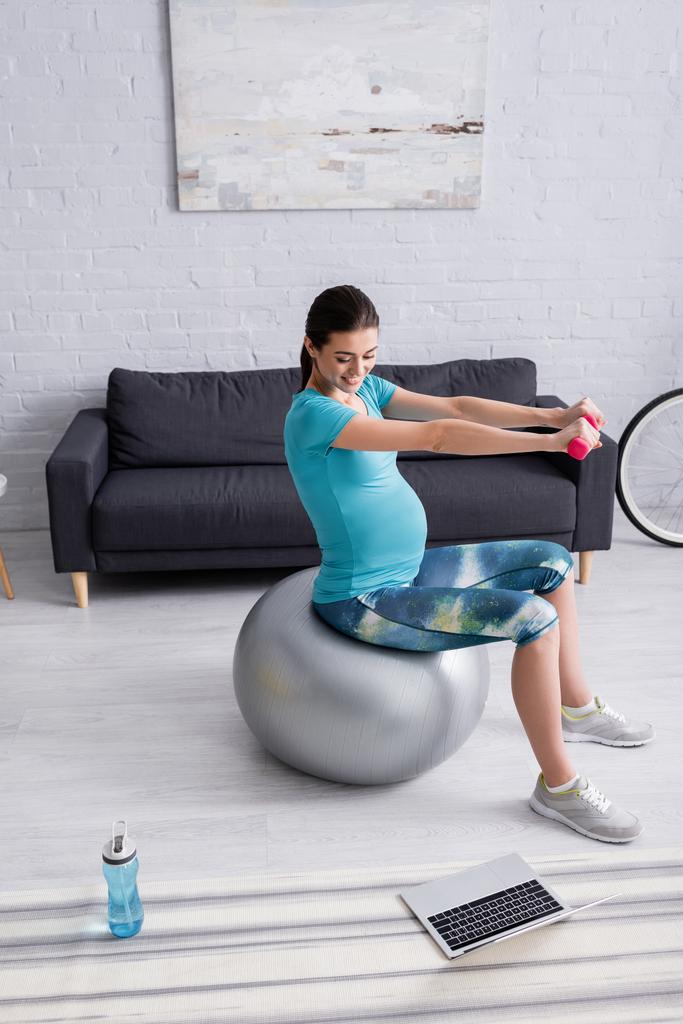 улыбающаяся беременная женщина тренируется на фитнес-мяч с гантелями возле ноутбука - Фото, изображение