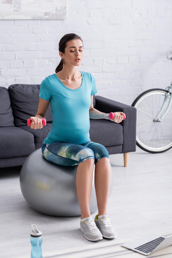 deportista embarazada haciendo ejercicio en la pelota de fitness con pesas cerca de la botella de deportes y portátil  - Foto, imagen