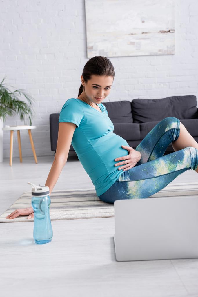 スポーツウェアを着た妊婦がフィットネスマットに座ってリビングのノートパソコンを見て  - 写真・画像