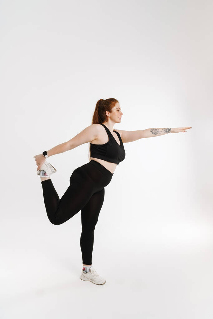 Souriant jeune femme de remise en forme plus la taille en vêtements de sport debout sur fond blanc faire des exercices d'étirement - Photo, image