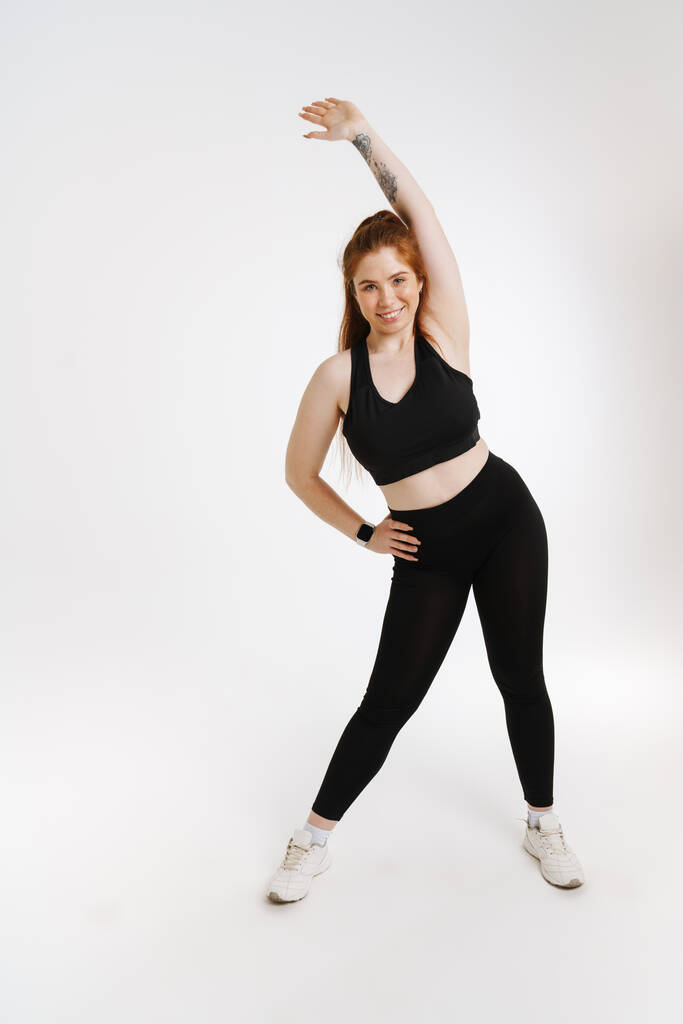 Usmívající se mladý plus velikost fitness žena ve sportovním oblečení stojící nad bílým pozadím dělá strečink cvičení - Fotografie, Obrázek