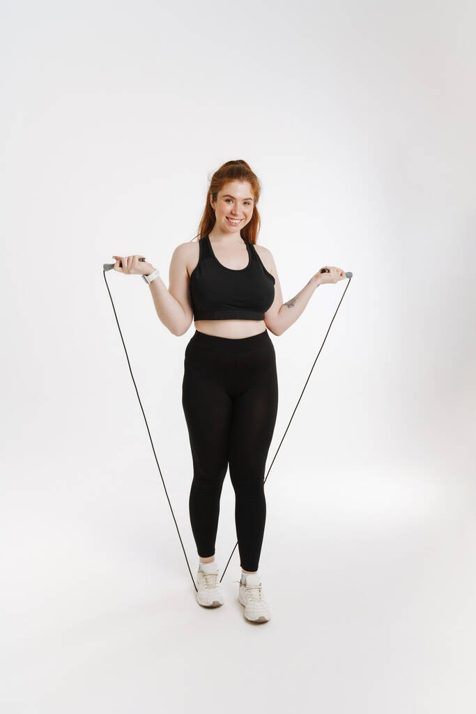 Usmívající se mladý plus velikost fitness žena ve sportovním oblečení stojící nad bílým pozadím cvičení s skákacím lanem plné délky - Fotografie, Obrázek