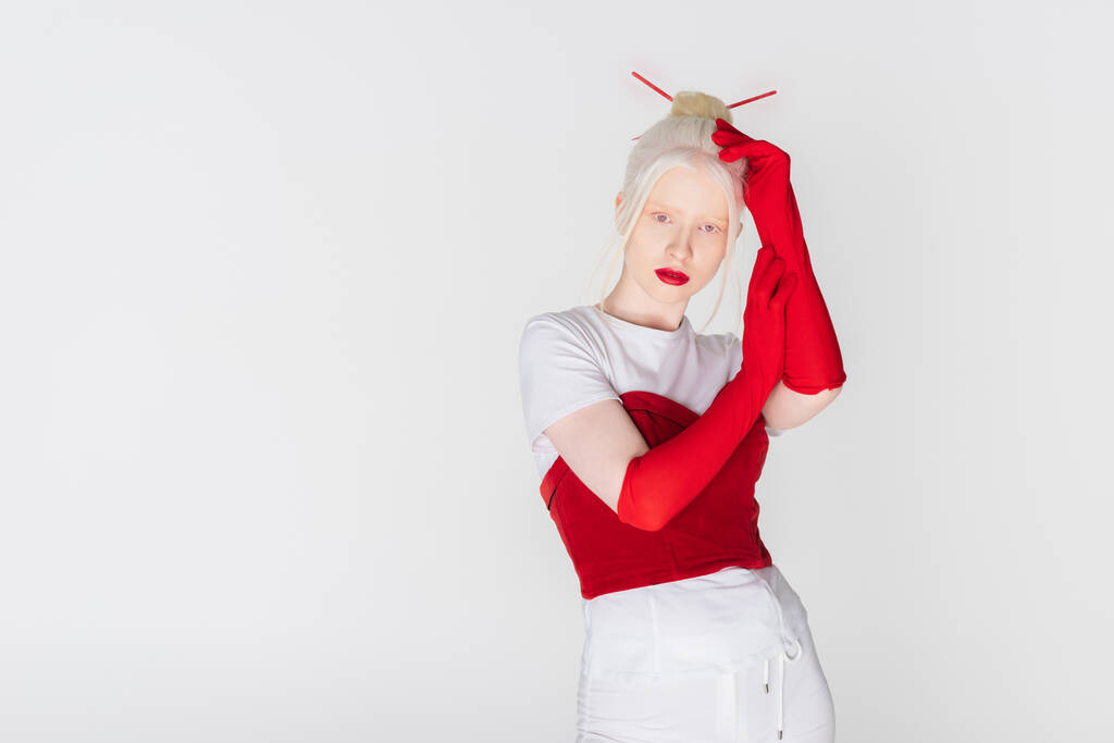 Modelo albino jovem em luvas vermelhas e varas de cabelo olhando para a câmera isolada no branco - Foto, Imagem