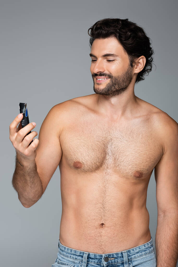 молодий без сорочки чоловік тримає електричну бритву і посміхається ізольовано на сірому
 - Фото, зображення
