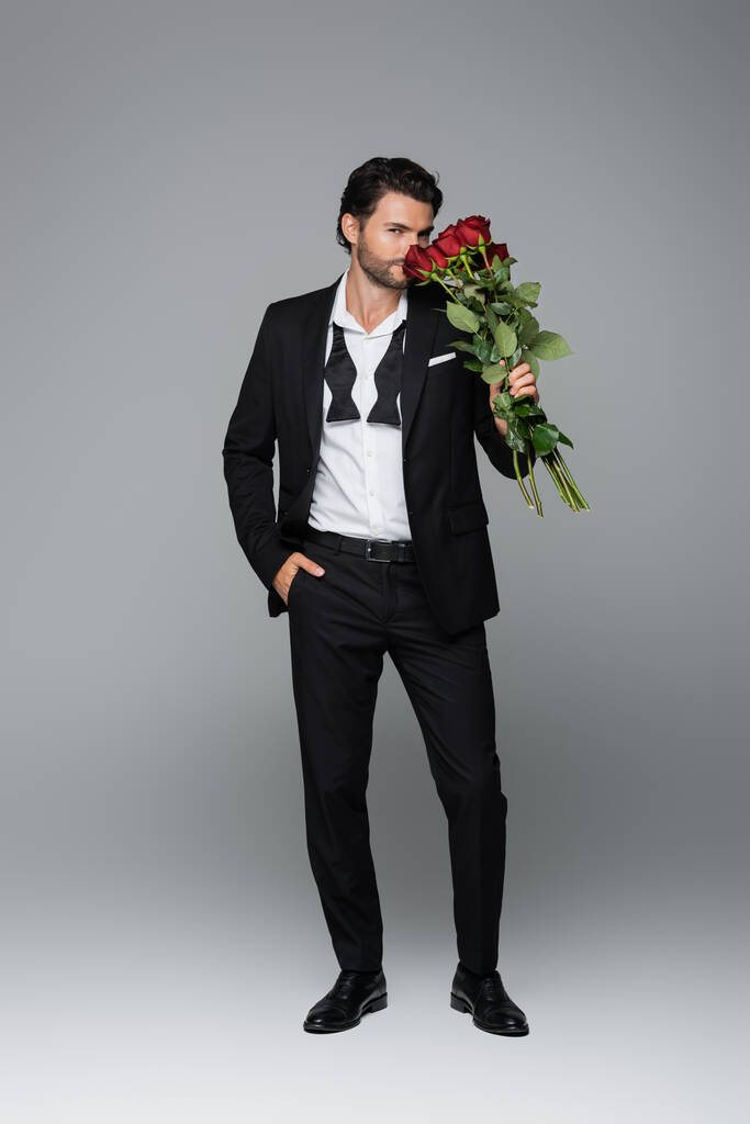 full length of bearded man in suit smelling red roses while standing with hand in pocket on grey - Photo, Image