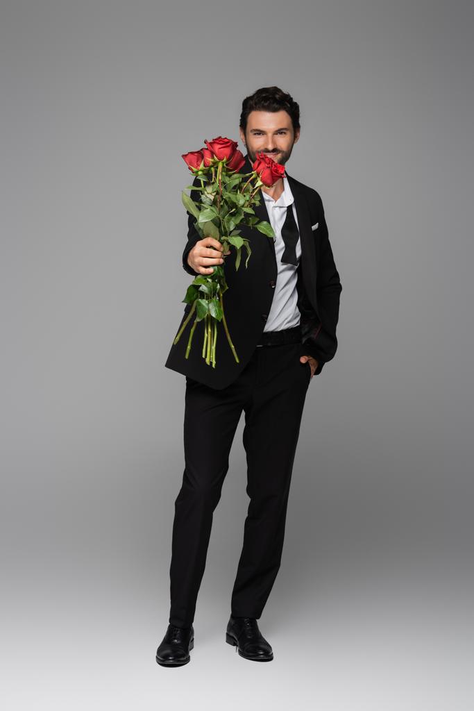 longitud completa de hombre feliz en traje sosteniendo rosas rojas mientras está de pie con la mano en el bolsillo en gris - Foto, imagen