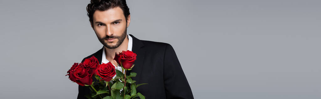 homme barbu en costume tenant bouquet de roses rouges tout en regardant la caméra isolée sur gris, bannière - Photo, image