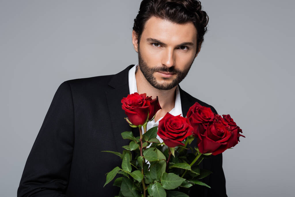 szakállas fiatalember öltönyben kezében csokor vörös rózsa, miközben néz kamera elszigetelt szürke - Fotó, kép