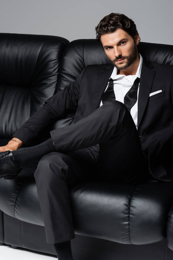 татуированный и бородатый мужчина в формальной одежде сидит на черном диване, изолированном на сером - Фото, изображение