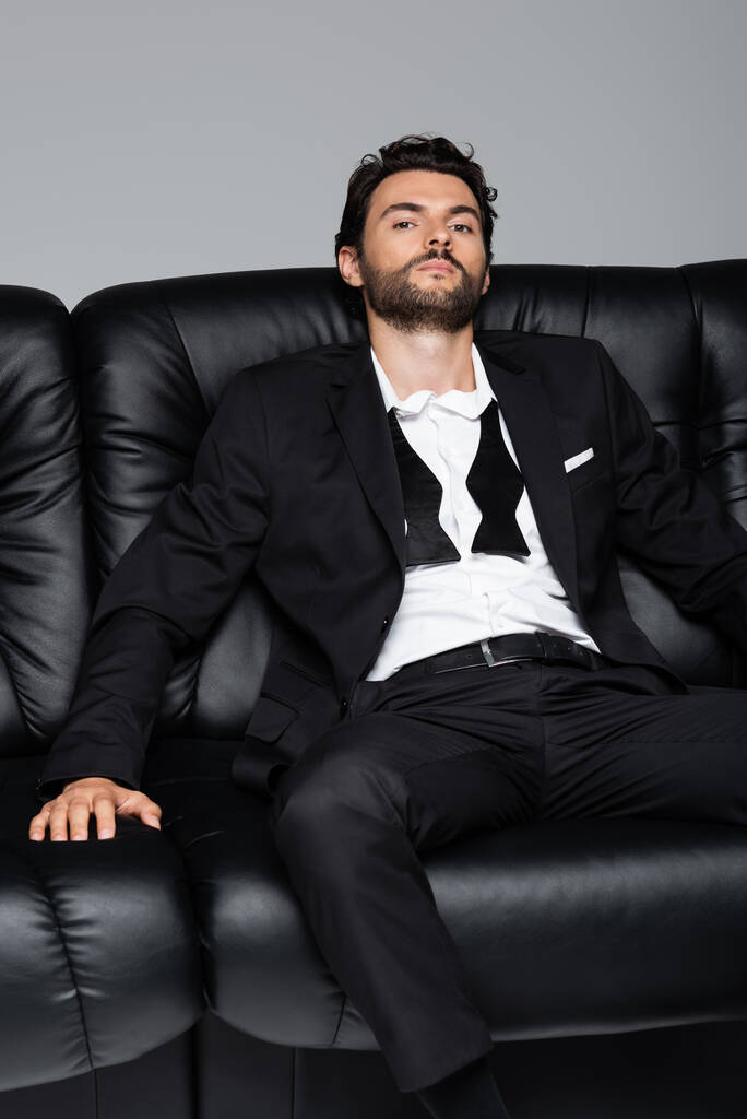 νεαρός γενειοφόρος άνδρας με επίσημη ένδυση κάθεται σε μαύρο καναπέ απομονωμένος σε γκρι - Φωτογραφία, εικόνα
