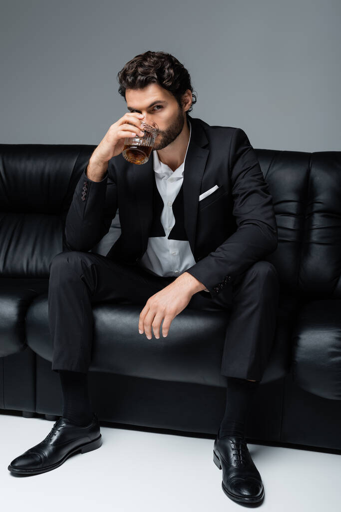 młody brodaty mężczyzna w formalnym ubraniu siedzi na czarnej sofie i pije whisky na szarości - Zdjęcie, obraz