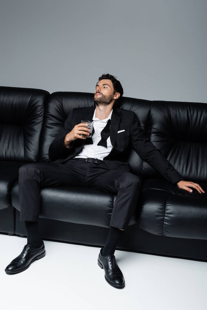 χαλαρός άνδρας με επίσημη ένδυση κάθεται σε μαύρο καναπέ και κρατώντας ένα ποτήρι ουίσκι σε γκρι - Φωτογραφία, εικόνα