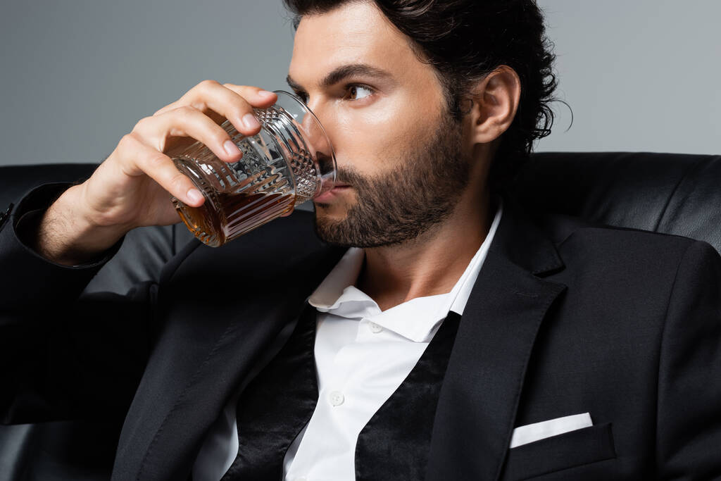 barbudo con cabello ondulado bebiendo whisky aislado en gris - Foto, Imagen