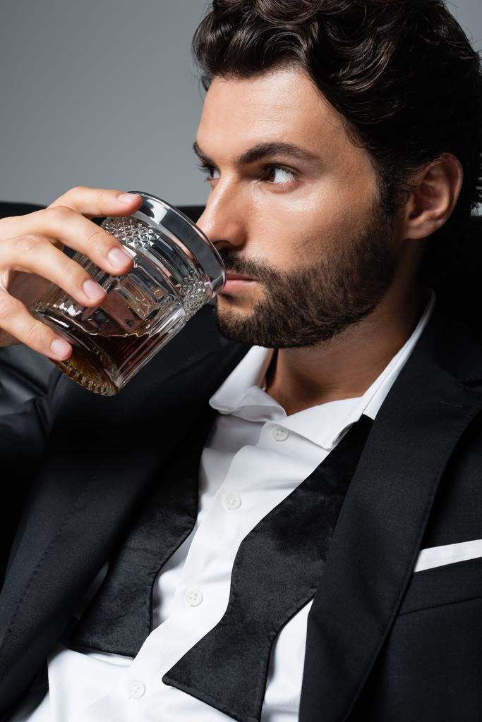 uomo barbuto con capelli ondulati bere brandy isolato su grigio - Foto, immagini