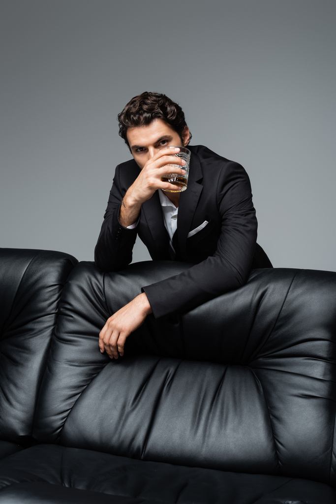 άνδρας με κοστούμι στέκεται κοντά σε μαύρο καναπέ και κρατώντας ένα ποτήρι ουίσκι απομονωμένο σε γκρι - Φωτογραφία, εικόνα