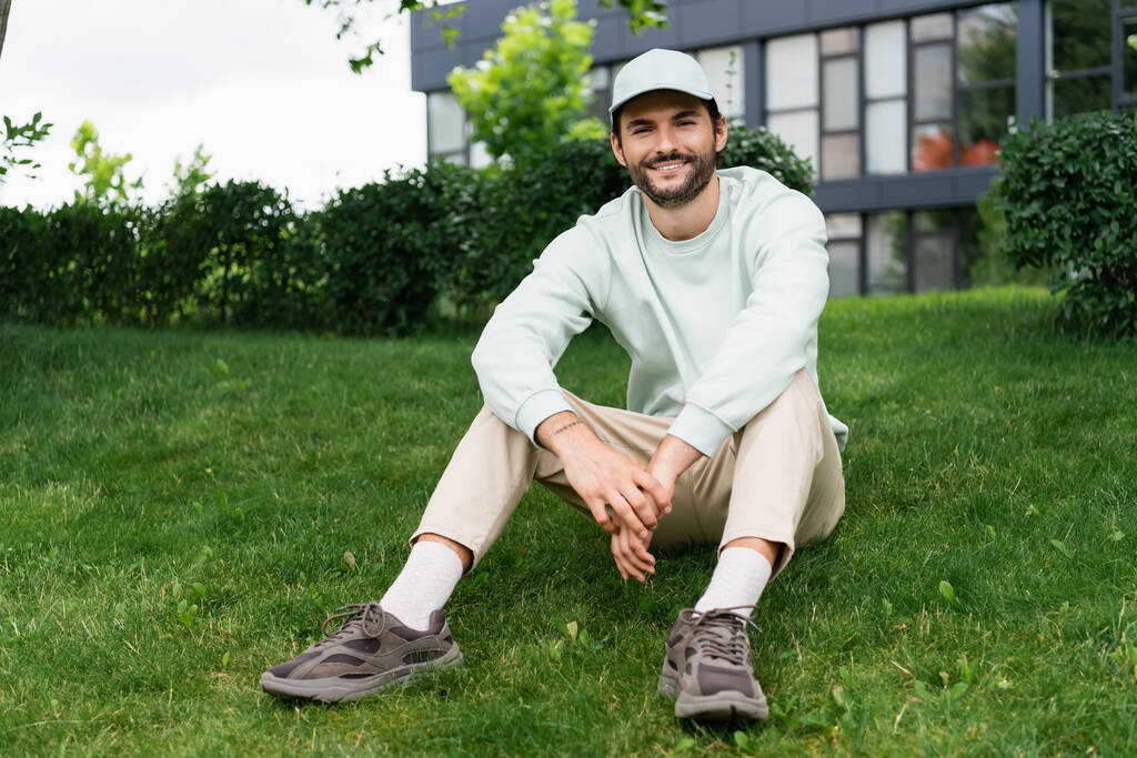 comprimento total de homem feliz em boné sorrindo enquanto sentado no gramado verde - Foto, Imagem