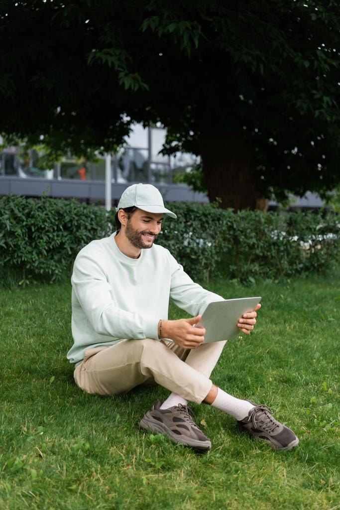 comprimento total de freelancer alegre usando laptop enquanto sentado com pernas cruzadas no gramado  - Foto, Imagem