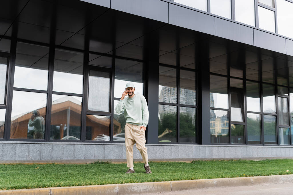 comprimento total de homem alegre de pé com a mão no bolso e falando no celular perto do edifício moderno  - Foto, Imagem