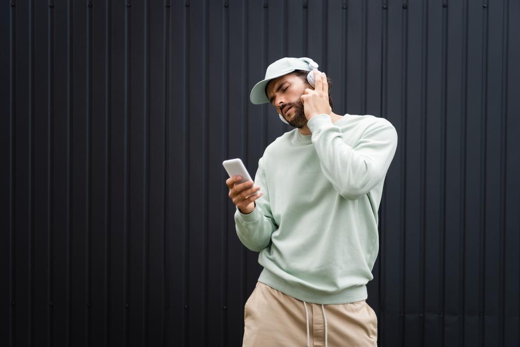 bärtiger Mann mit Baseballmütze hört Musik in drahtlosen Kopfhörern in der Nähe von Metallzaun - Foto, Bild