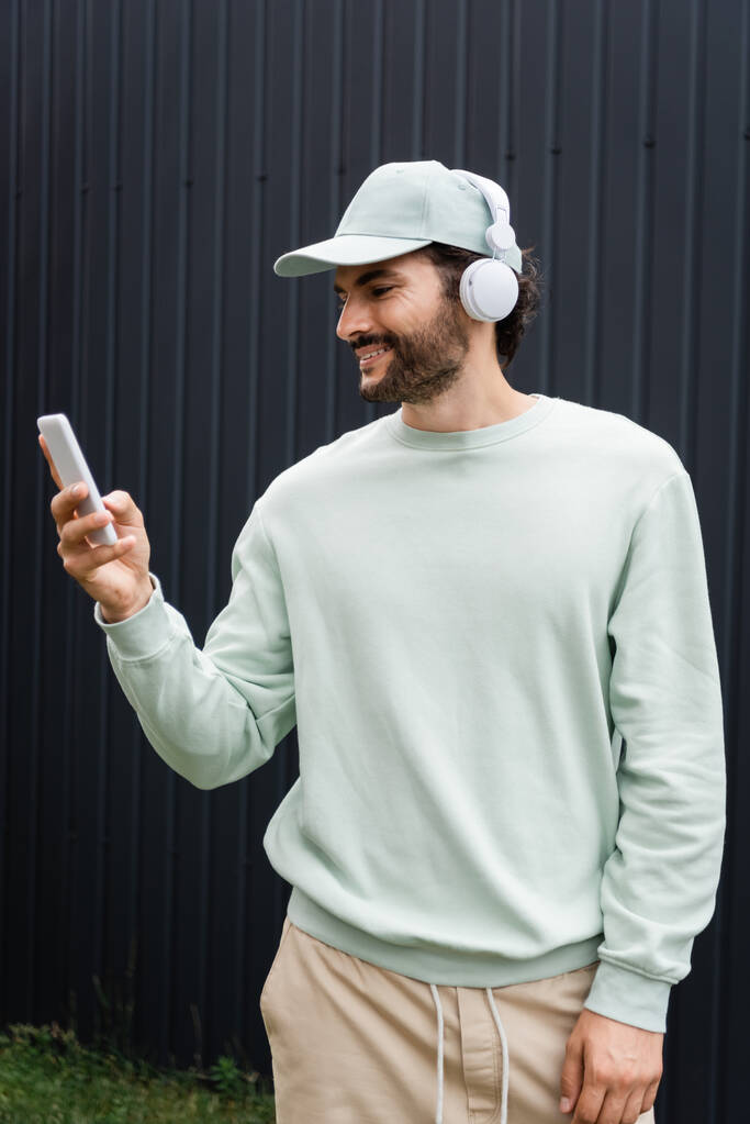 pozytywny człowiek w czapce słuchanie muzyki w słuchawkach bezprzewodowych i trzymanie telefonu komórkowego w pobliżu metalowego ogrodzenia - Zdjęcie, obraz