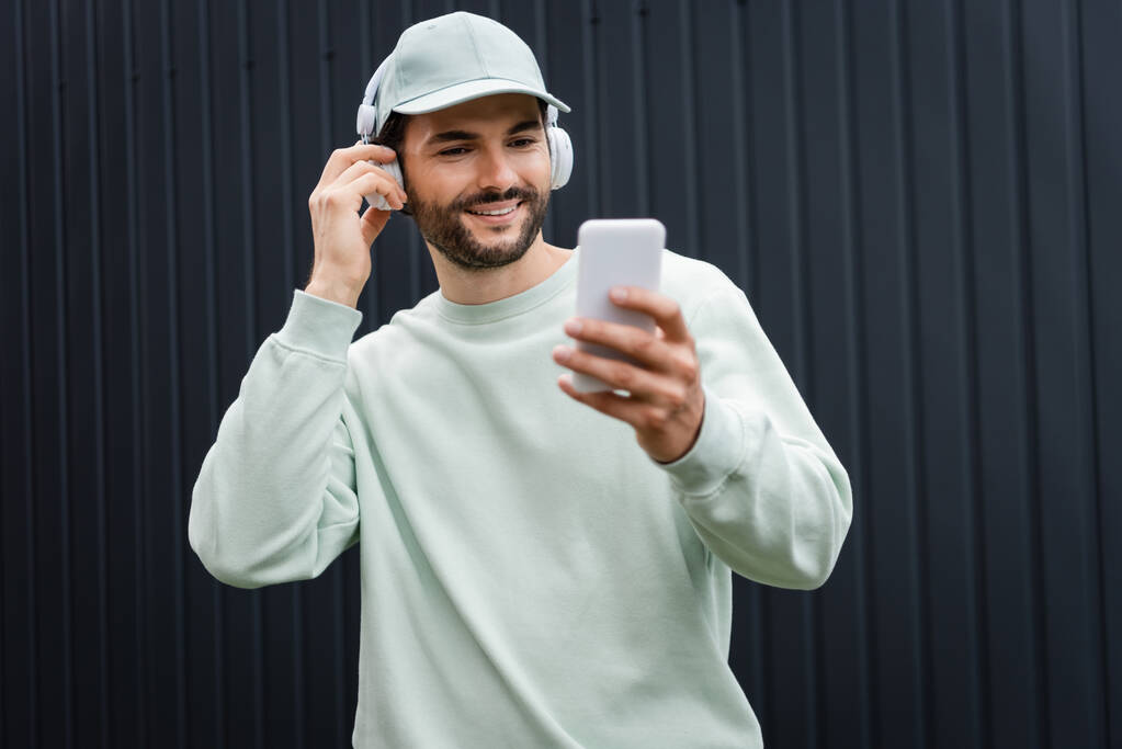 pozytywny człowiek w czapce słuchanie muzyki w słuchawkach bezprzewodowych i za pomocą telefonu komórkowego w pobliżu metalowego ogrodzenia - Zdjęcie, obraz