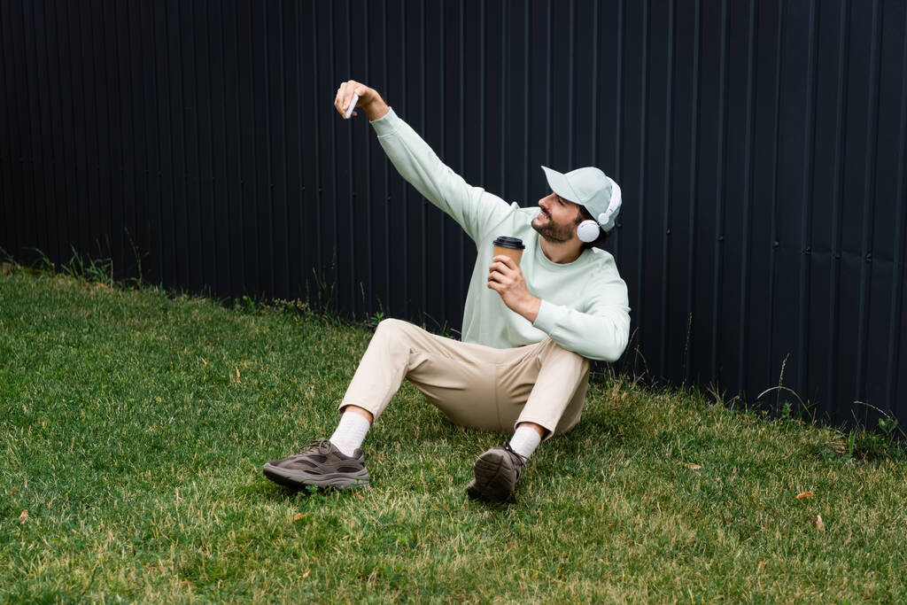 szczęśliwy człowiek w bezprzewodowych słuchawkach robi selfie siedząc na zielonym trawniku z papierową filiżanką  - Zdjęcie, obraz