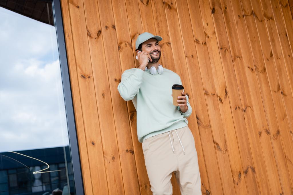 Kapüşonlu mutlu adam elinde kahve tutarken binanın yanındaki akıllı telefondan konuşuyor.  - Fotoğraf, Görsel