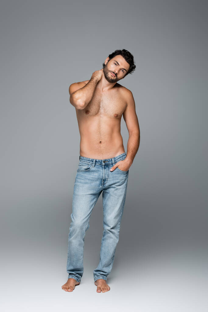 full length of good-looking and muscular man in blue jeans posing with hand in pocket on grey - Photo, Image