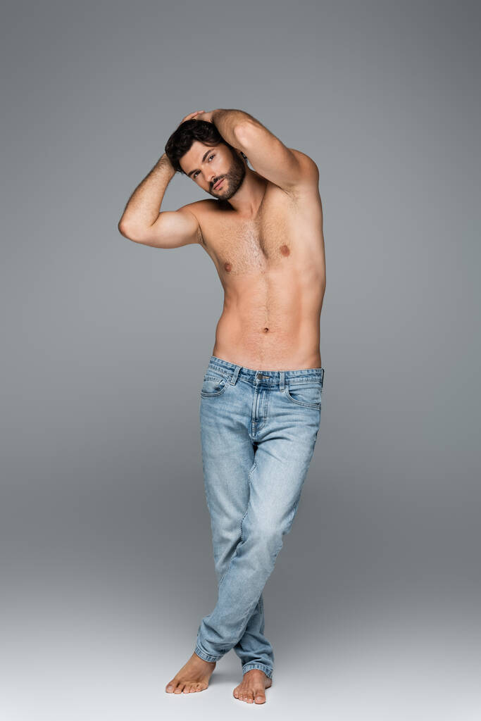 comprimento total de homem bonito e musculoso em jeans olhando para a câmera enquanto posando em cinza - Foto, Imagem