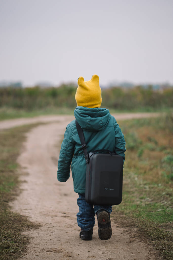Маленький кавказский мальчик несет большую сумку с беспилотником в поле, вид сзади. - Фото, изображение